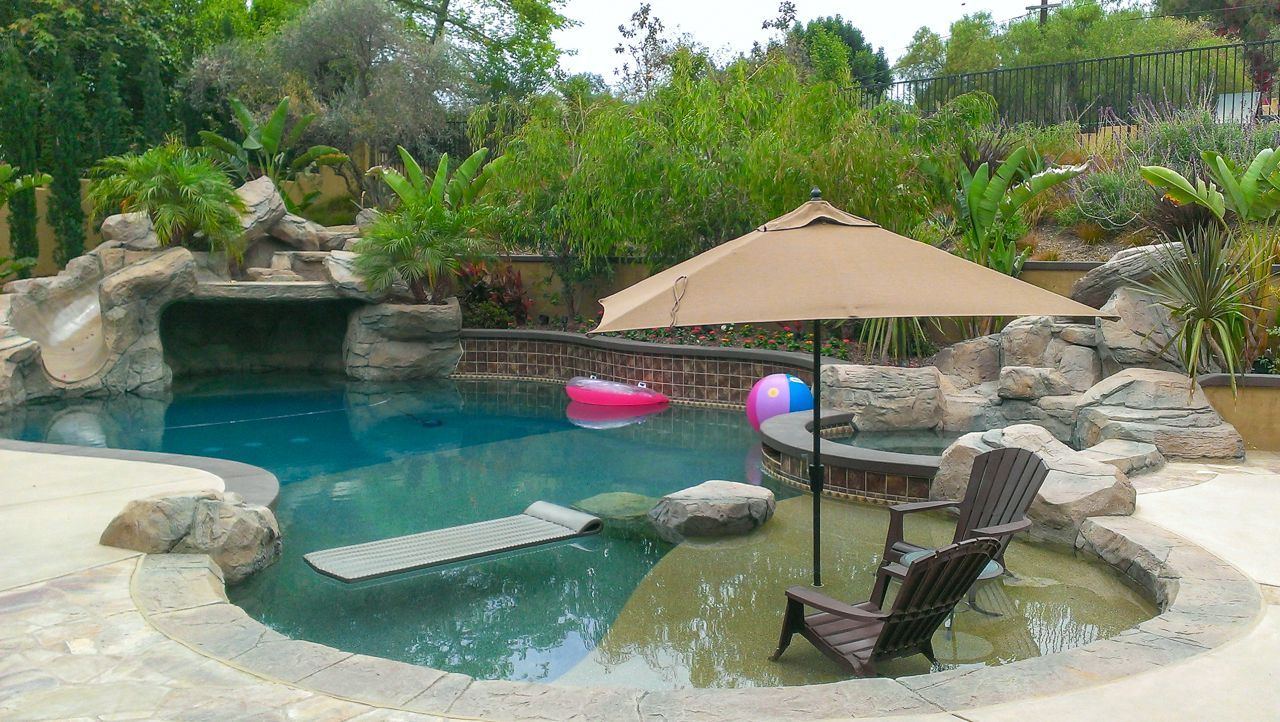 Swimming pool with rocks, umbrella, and a lounge chair in a sunny backyard setting.