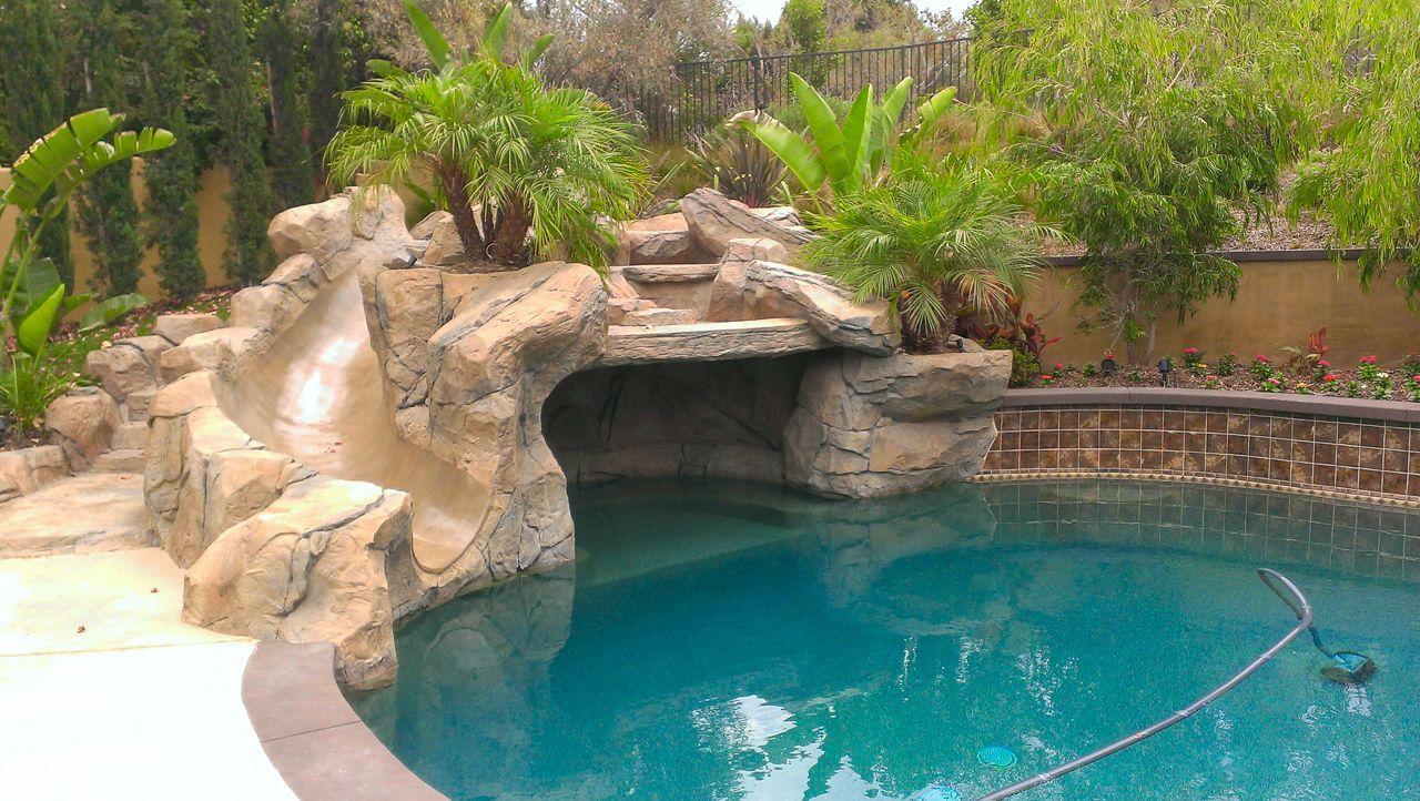 Swimming pool with rock waterfall, slide, and lush greenery.