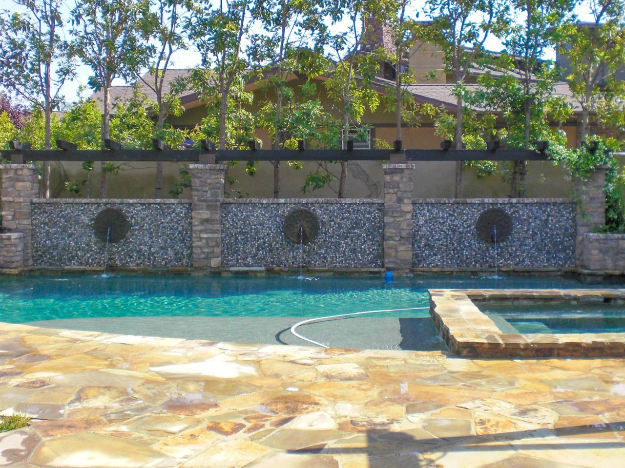 Swimming pool with water feature wall, stone patio, and trees in background.
