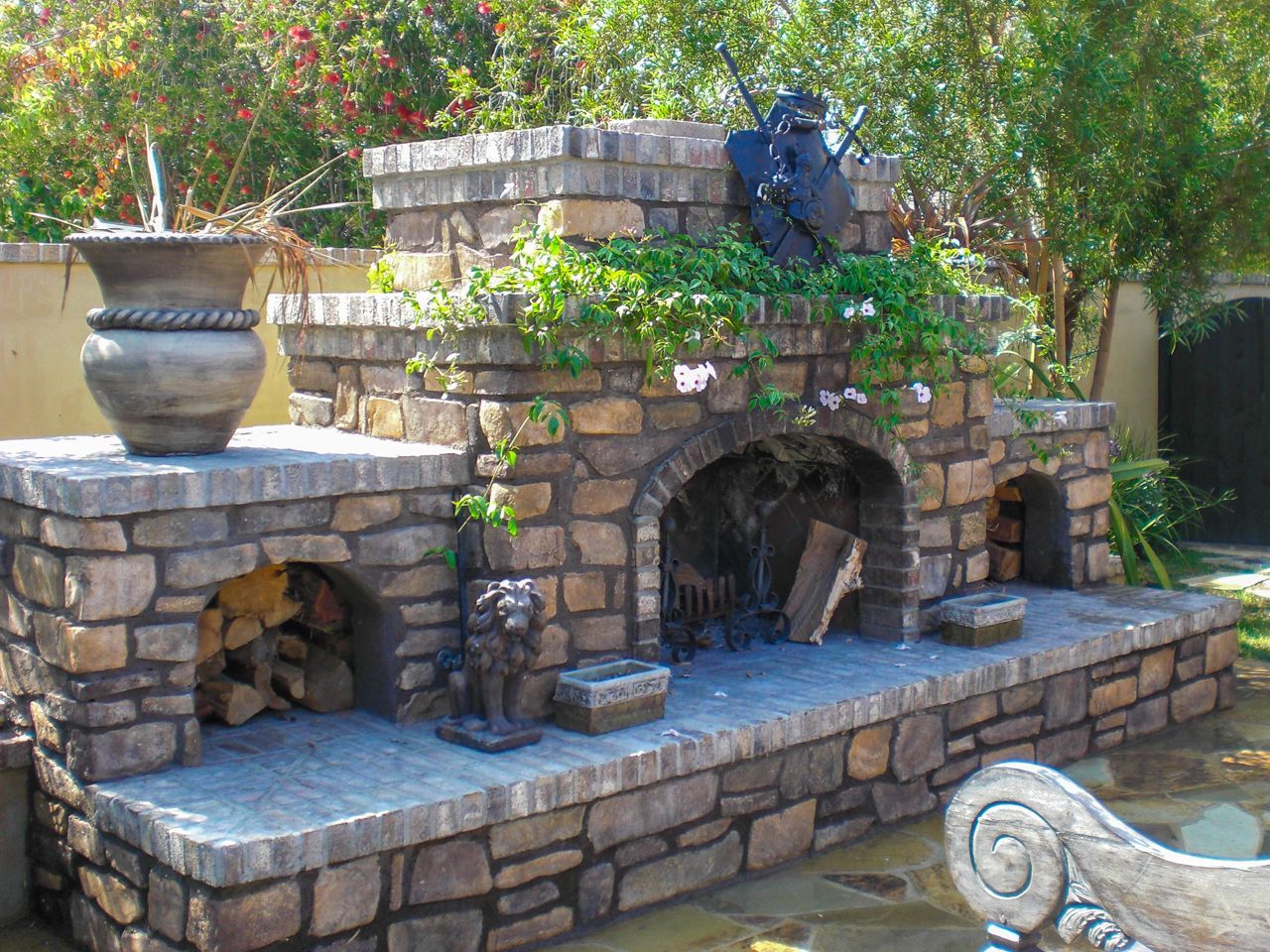 Stone outdoor fireplace with a wood storage area, decorative lion statues, and climbing greenery.