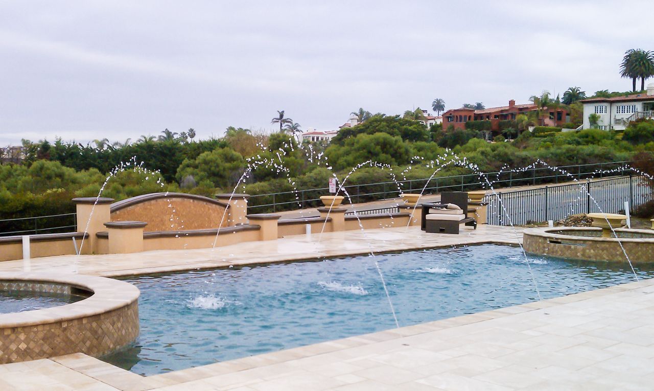 Swimming pool with water fountains and a surrounding patio, against a background of trees and houses under a cloudy sky.