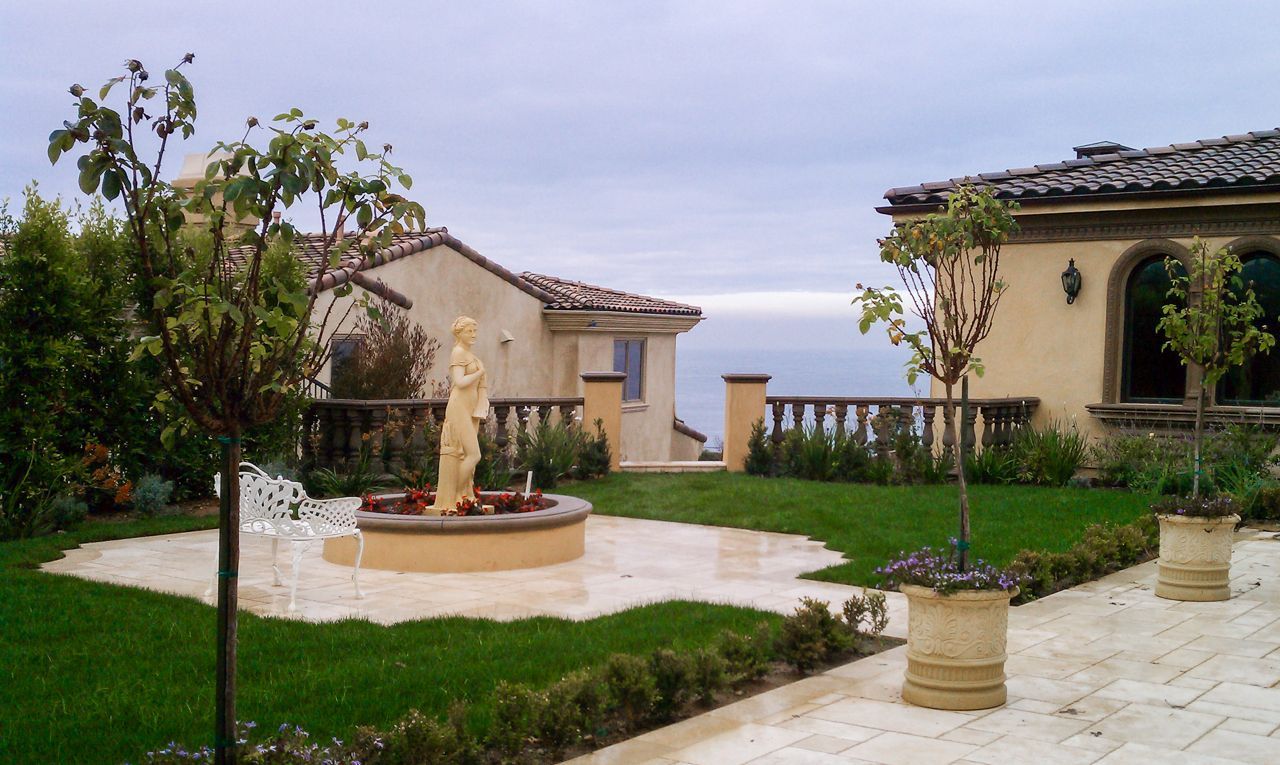 Exterior of house with fountain, lawn, and ocean view on a cloudy day.