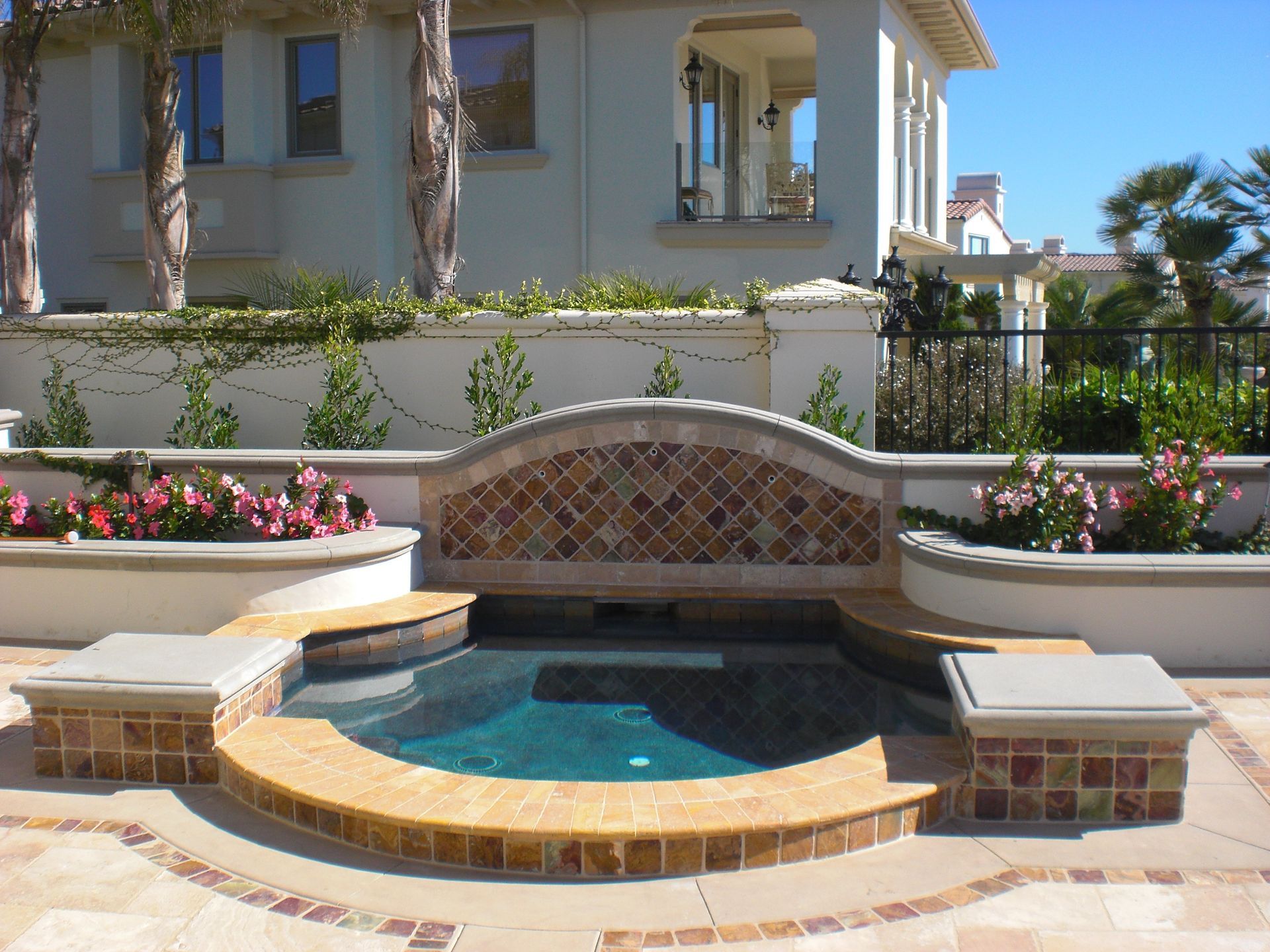 Outdoor spa with mosaic tile accents, surrounded by a patio and a cream-colored building.