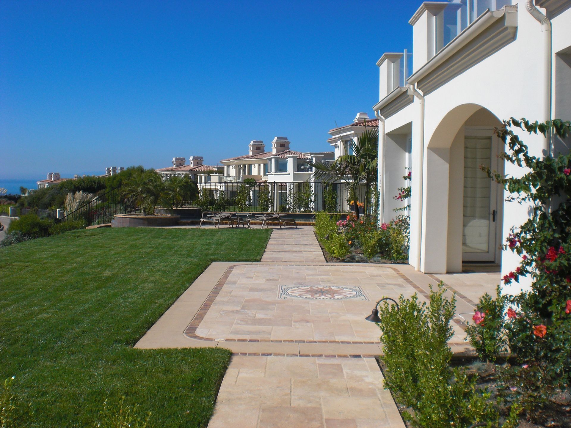 A luxurious white house with a paved pathway and green lawn overlooking the ocean under a clear blue sky.