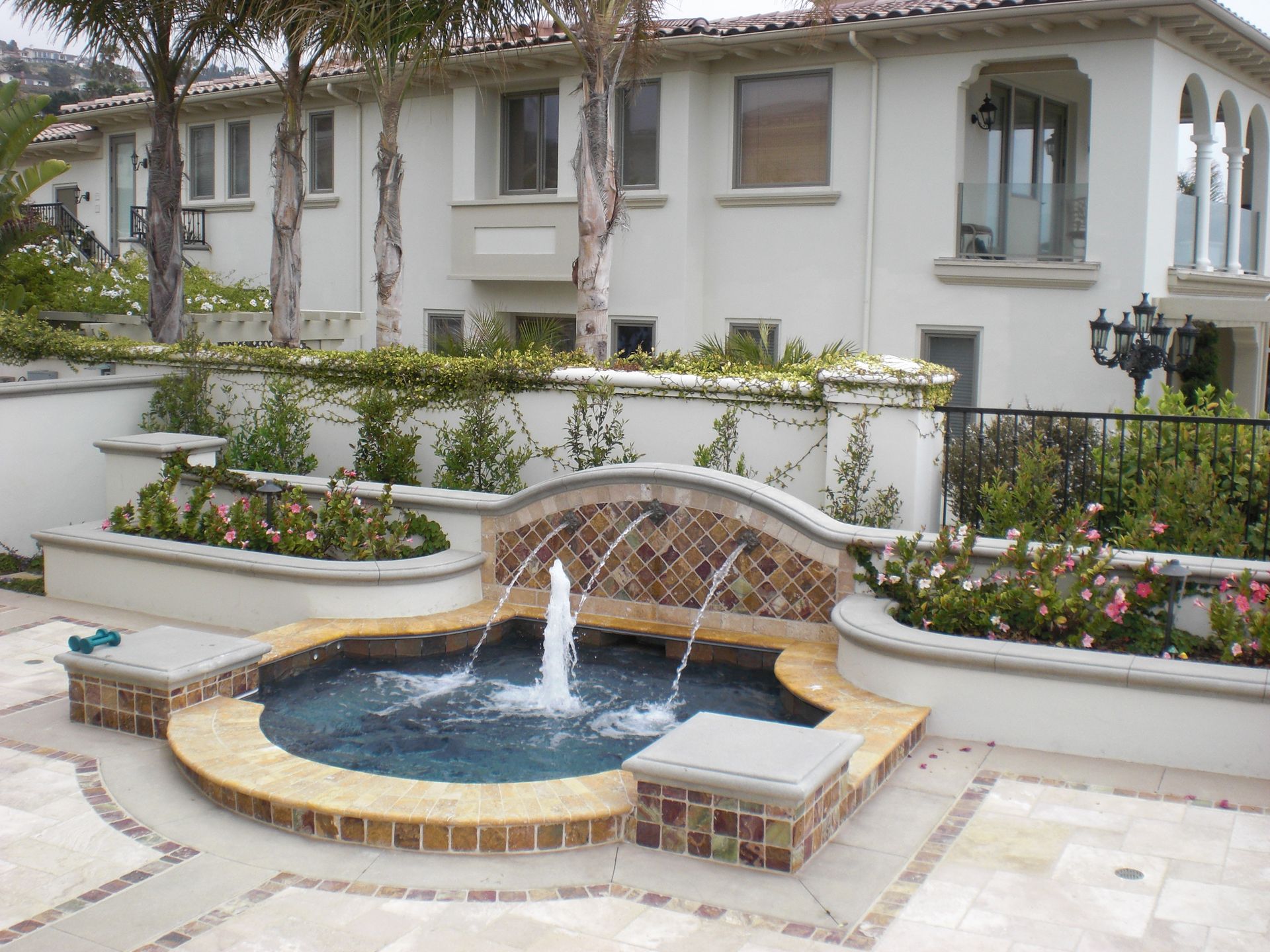 Fountain with decorative tile, surrounded by a patio and a large white house.