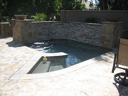 Stone-lined outdoor spa with steps, surrounded by stone patio and pillars topped with bowls. A rubber duck floats in the spa.