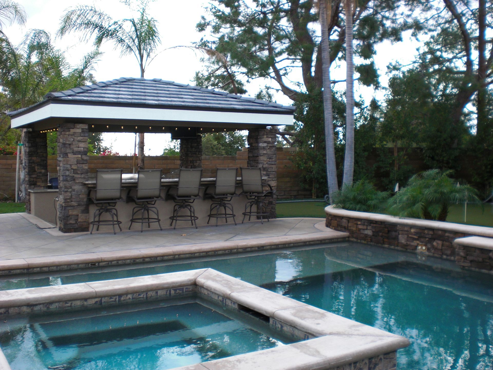 Outdoor bar with seating next to a pool. Brown stone columns and a tiled roof.
