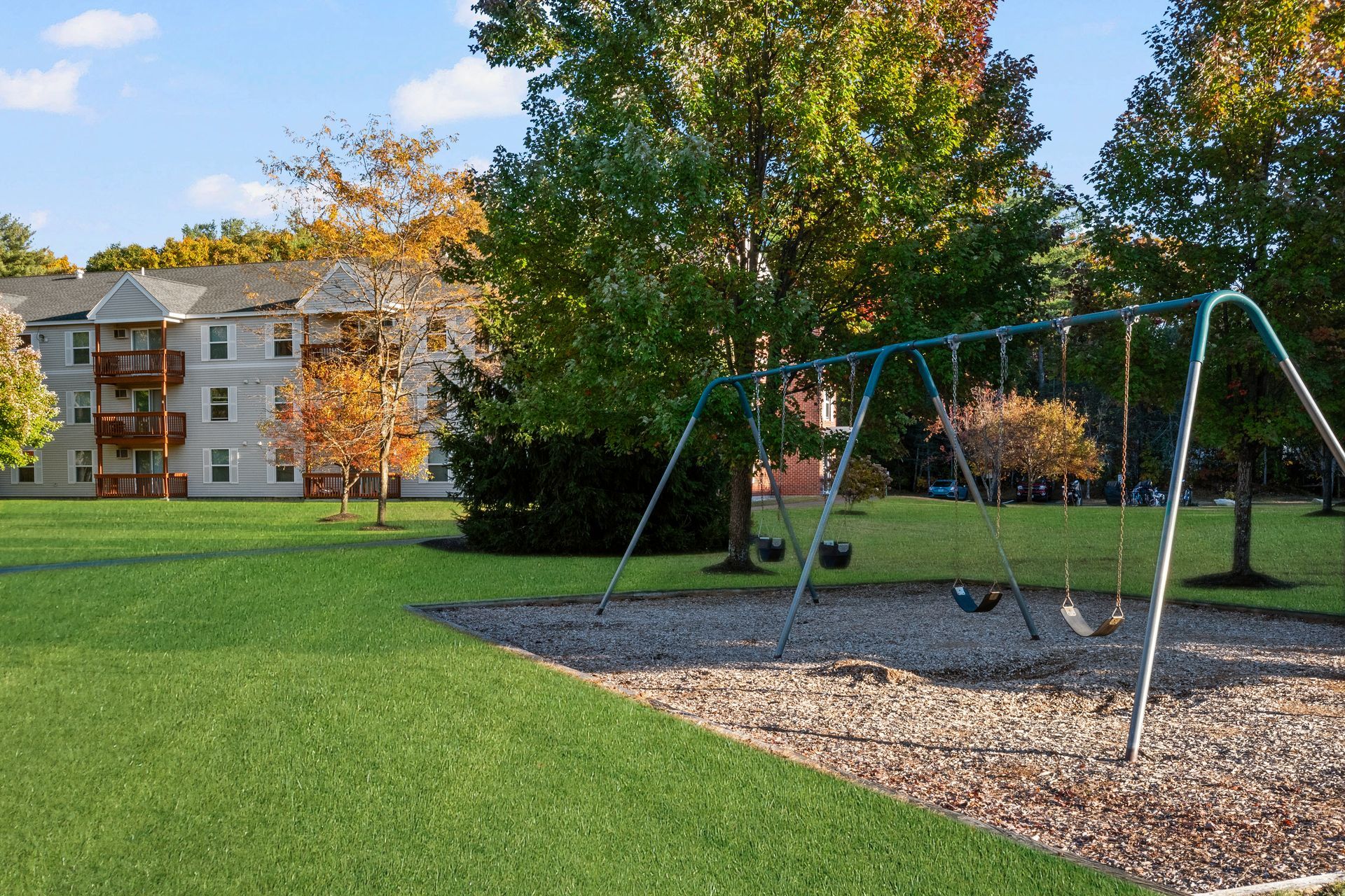 Apartment complex with green lawn, playground swing, trees, and blue sky at Penacook Place Apartments in Concord, NH.