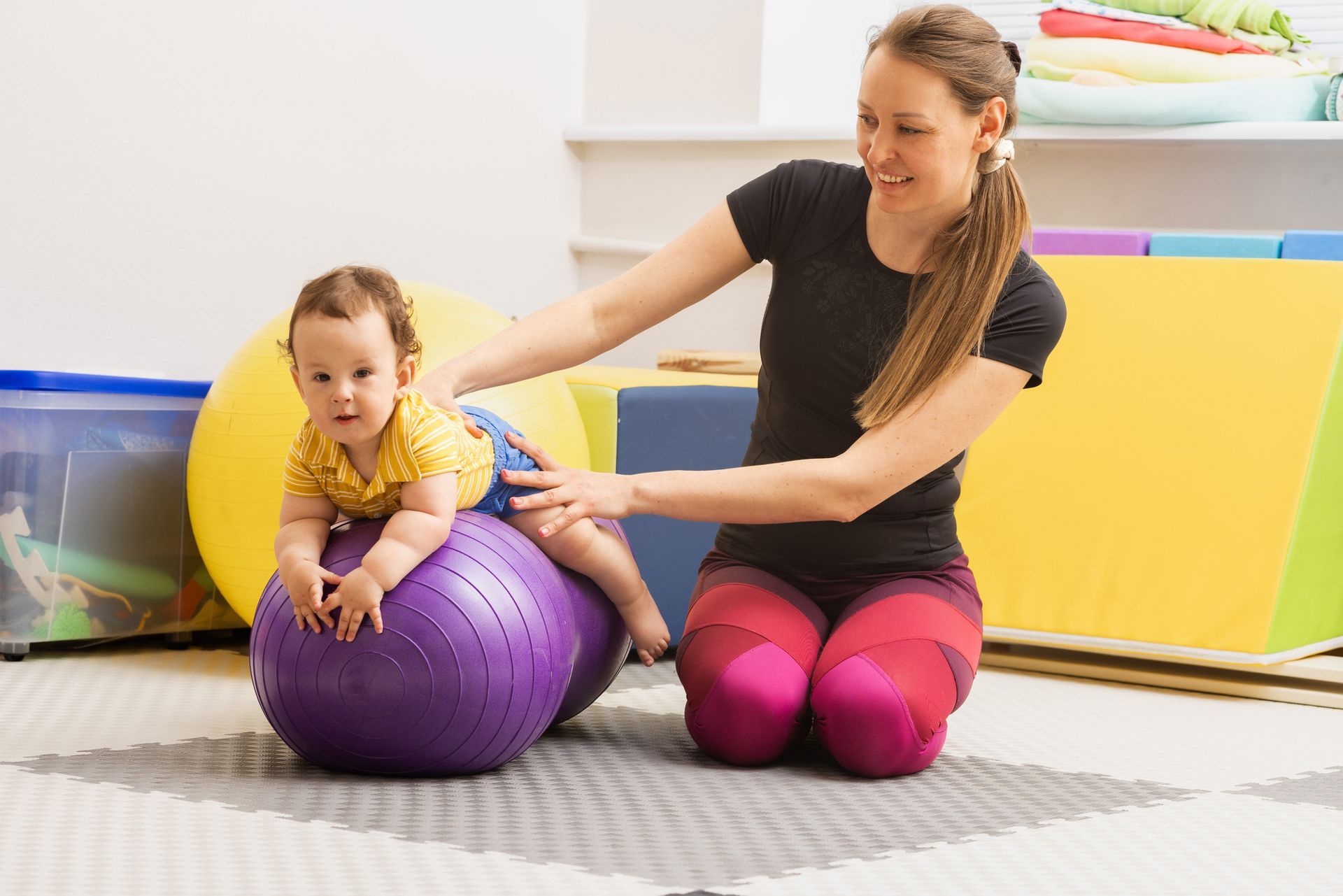 A woman is sitting on the floor with a baby on a purple exercise ball.