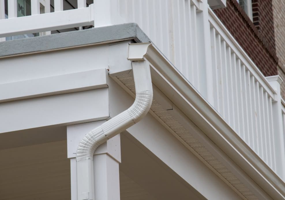 White gutter downspout on a house corner with a brick wall and porch railing above