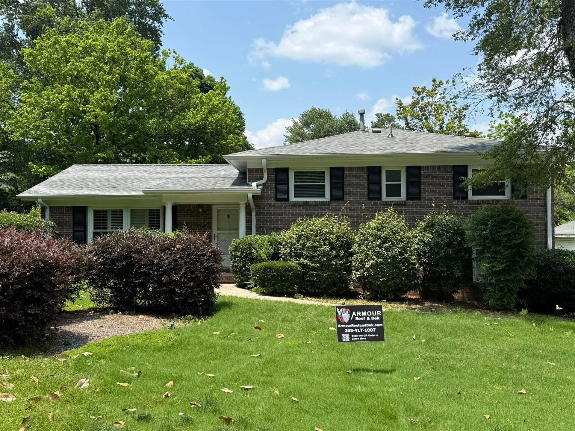 Suburban house with brick and white siding, green lawn, shrubs, and a small sign in front
