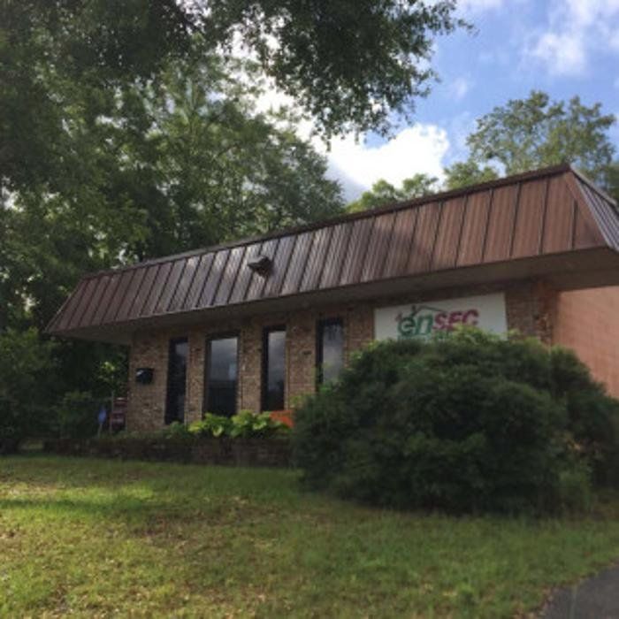 Small brick building with brown metal roof, surrounded by trees and shrubs on a grassy lawn.
