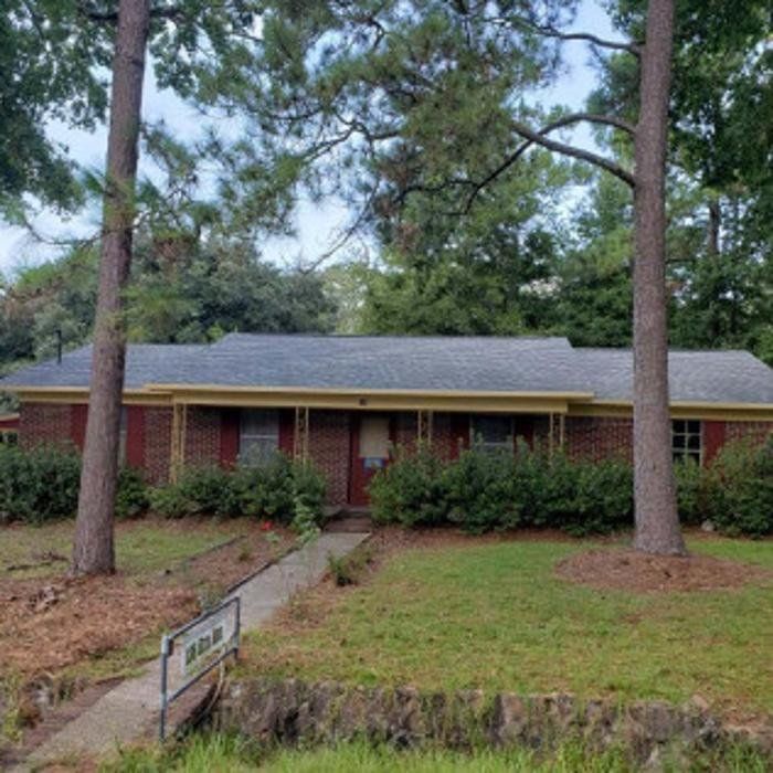 Single-story brick house with a front lawn, tall pine trees, and a walkway to the entrance