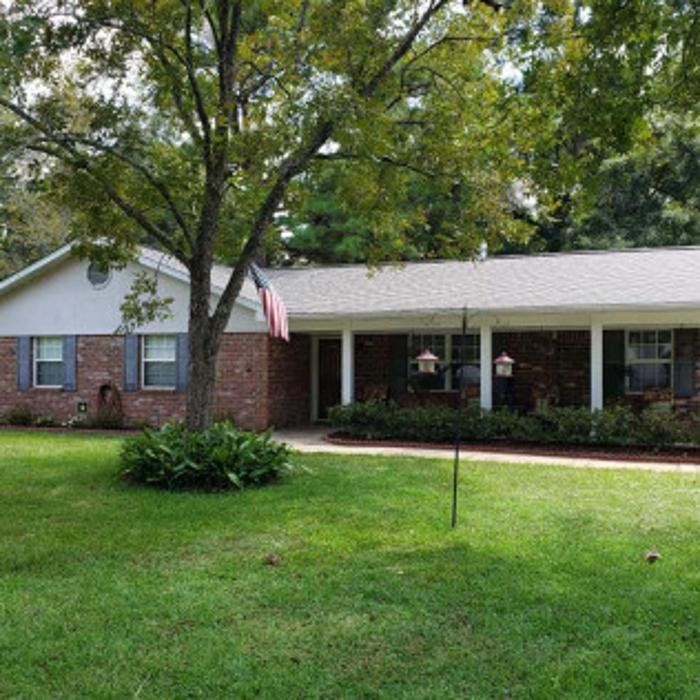 Single-story brick house with a large front lawn, trees, and a covered porch with an American flag.