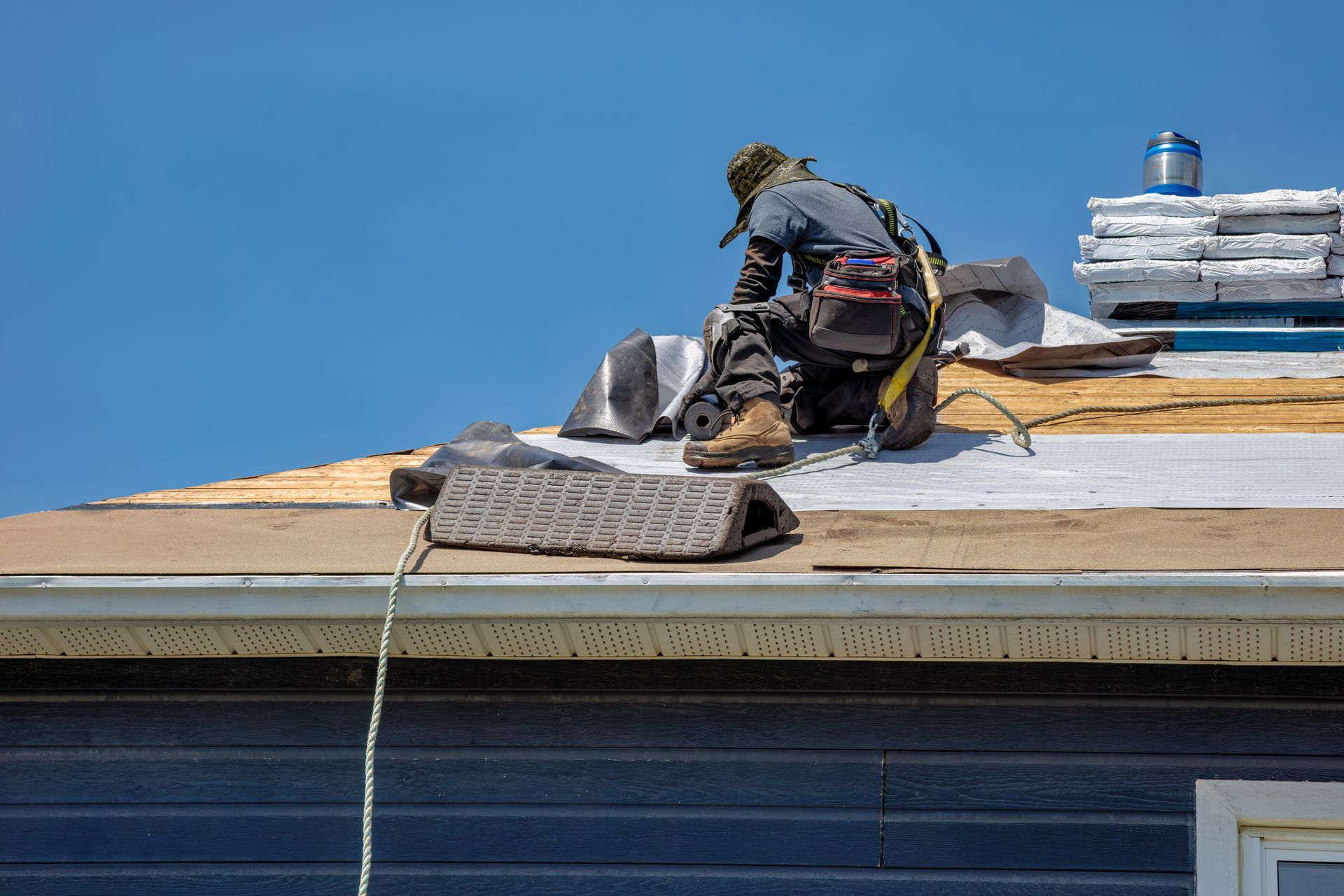 Worker on a roof installing materials beside a white vent under a clear blue sky