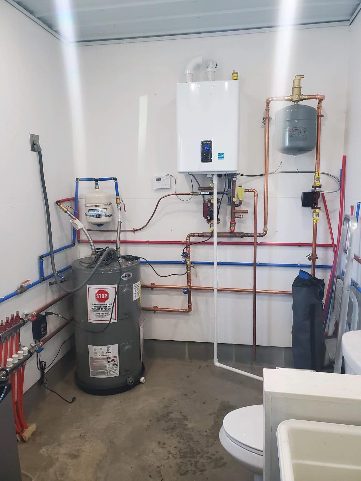 A water heater and related plumbing installed in a small, white-walled utility room.