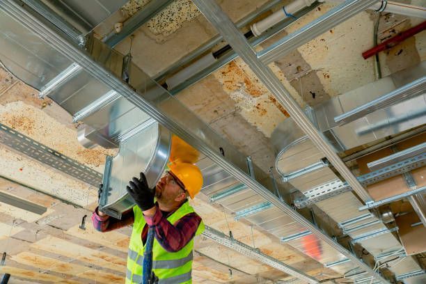 Construction worker in hard hat and vest installs ventilation ductwork in a building.