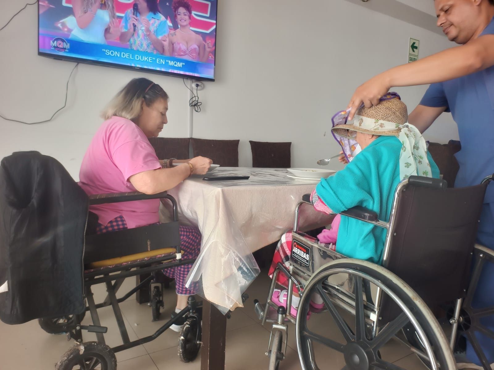 Cuidadora con uniforme azul sonriendo junto a una anciana en una luminosa sala de estar.