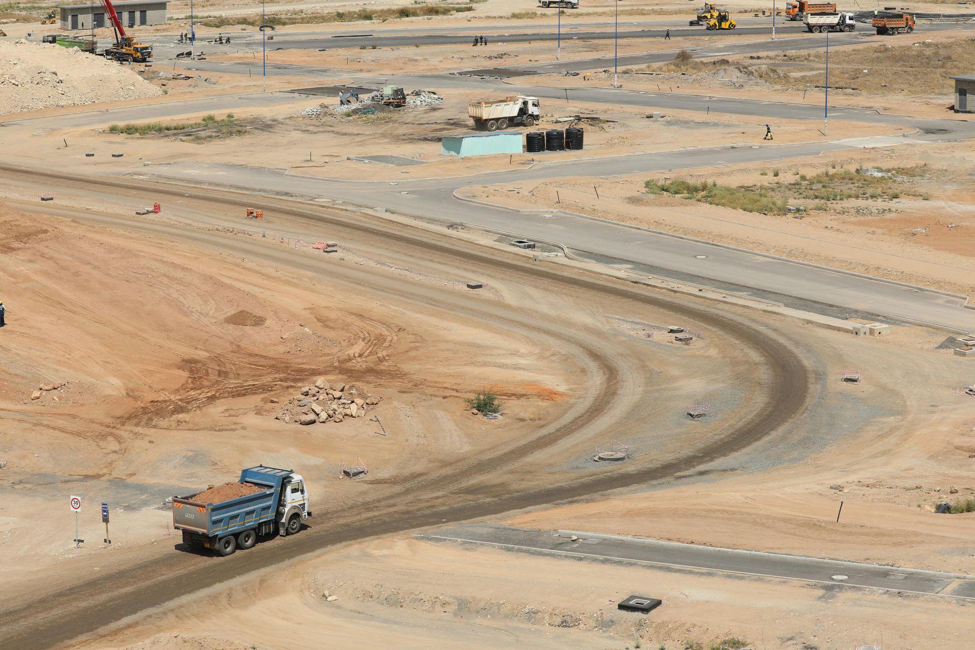 Curving desert road under construction with a blue truck and scattered workers in a sandy landscape