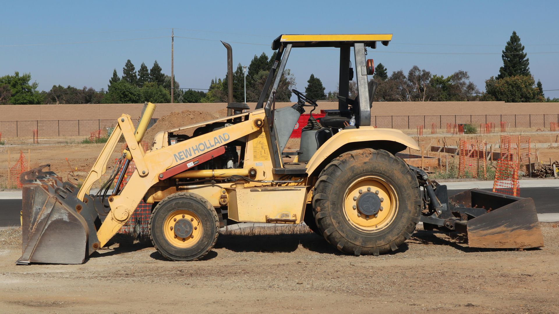 Yellow backhoe loader with front bucket and rear excavator arm on a dirt worksite.