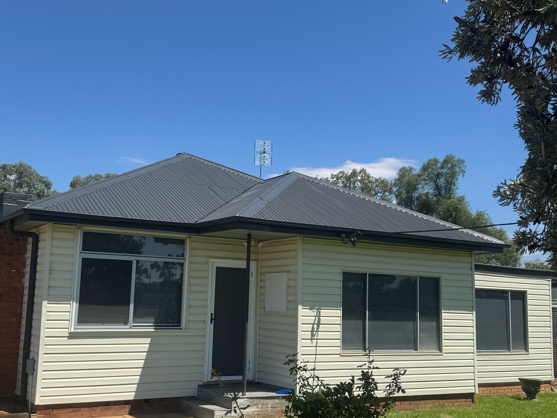 Single-story house with a dark roof, light siding, and a small front porch under a clear blue sky