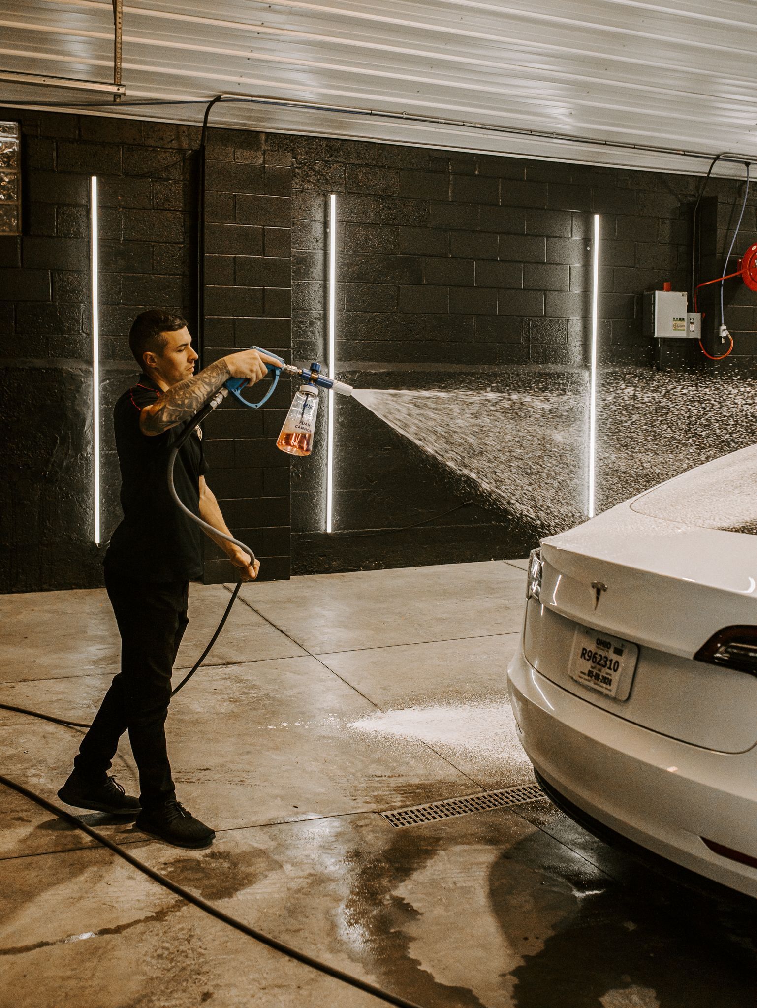 Person spraying water from a hose onto a white car in a dim garage-like space