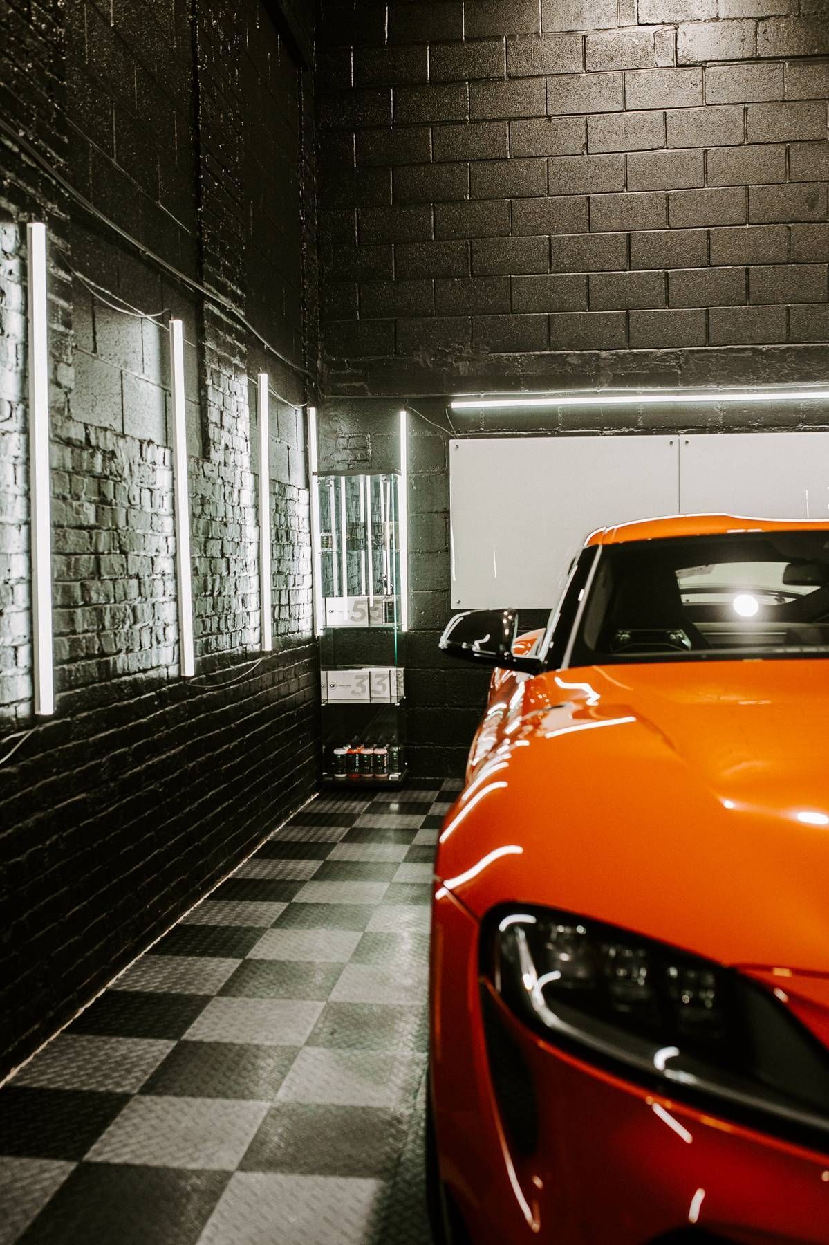 Orange sports car in a garage with black brick walls, checkered floor, and neon lights.