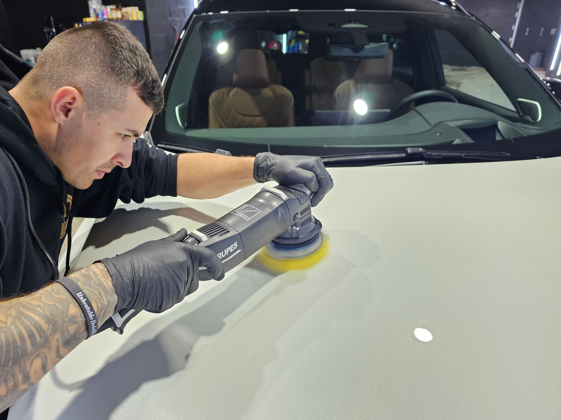 Detailing technician polishing a white car hood with a buffer in a garage