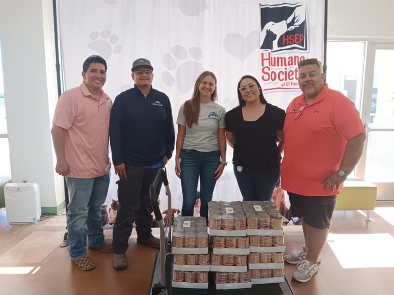Five people posing indoors beside stacked canned goods and a Human Society banner.