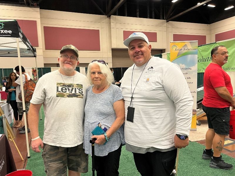 Three people posing at an indoor expo, two in white shirts and one in a gray shirt, with booths in the background