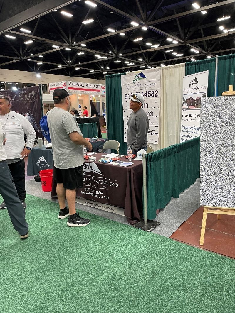 Trade show booth with three people talking beside a green curtain and display table on a convention floor.