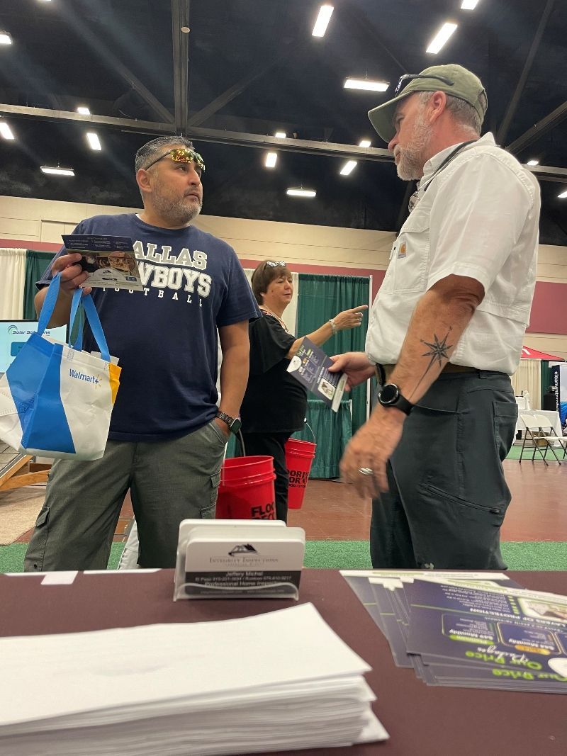 Two men talking at a vendor booth in a convention hall, with brochures and a table in the foreground.