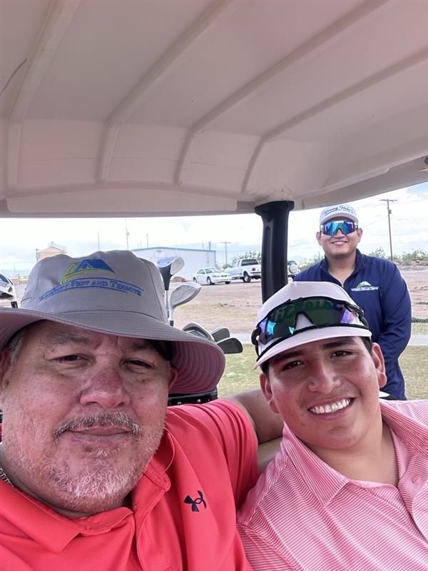 Three smiling people on a golf cart, wearing hats and sunglasses at a sunny outdoor event.
