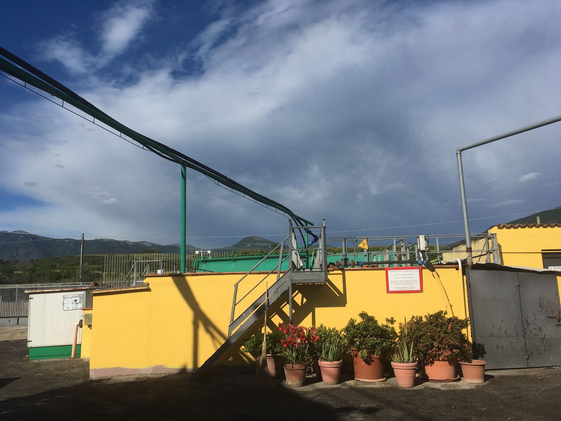 Edificio giallo con infrastrutture verdi, scale, piante in vaso. Cielo nuvoloso, montagne sullo sfondo.
