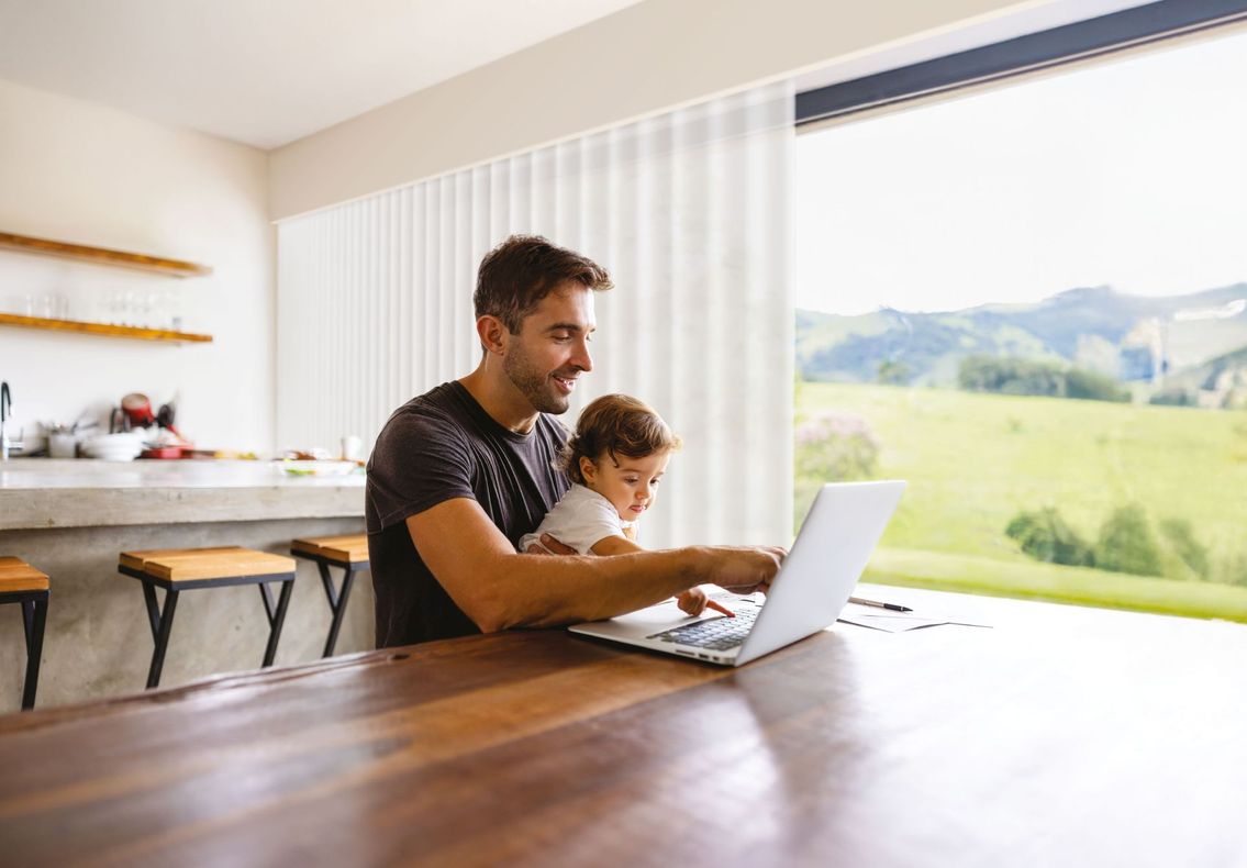 Un uomo e un bambino usano un computer portatile a un tavolo di legno accanto a una finestra luminosa, con verdi colline all'esterno.