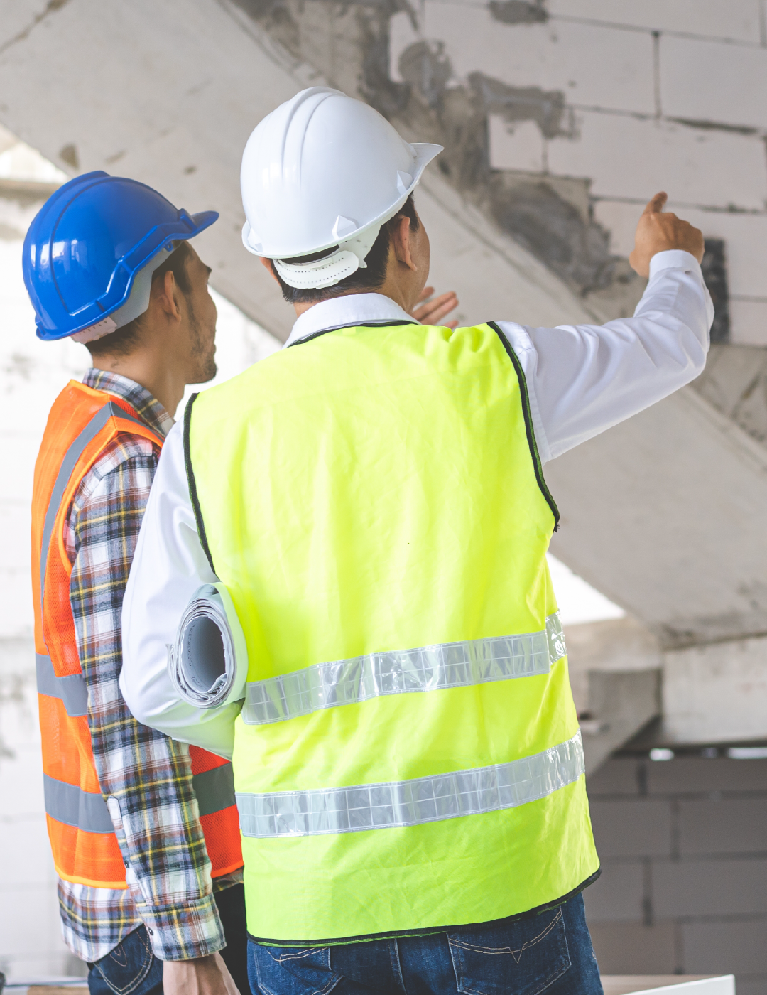 Two construction workers wearing hard hats and safety vests are looking at a brick wall.