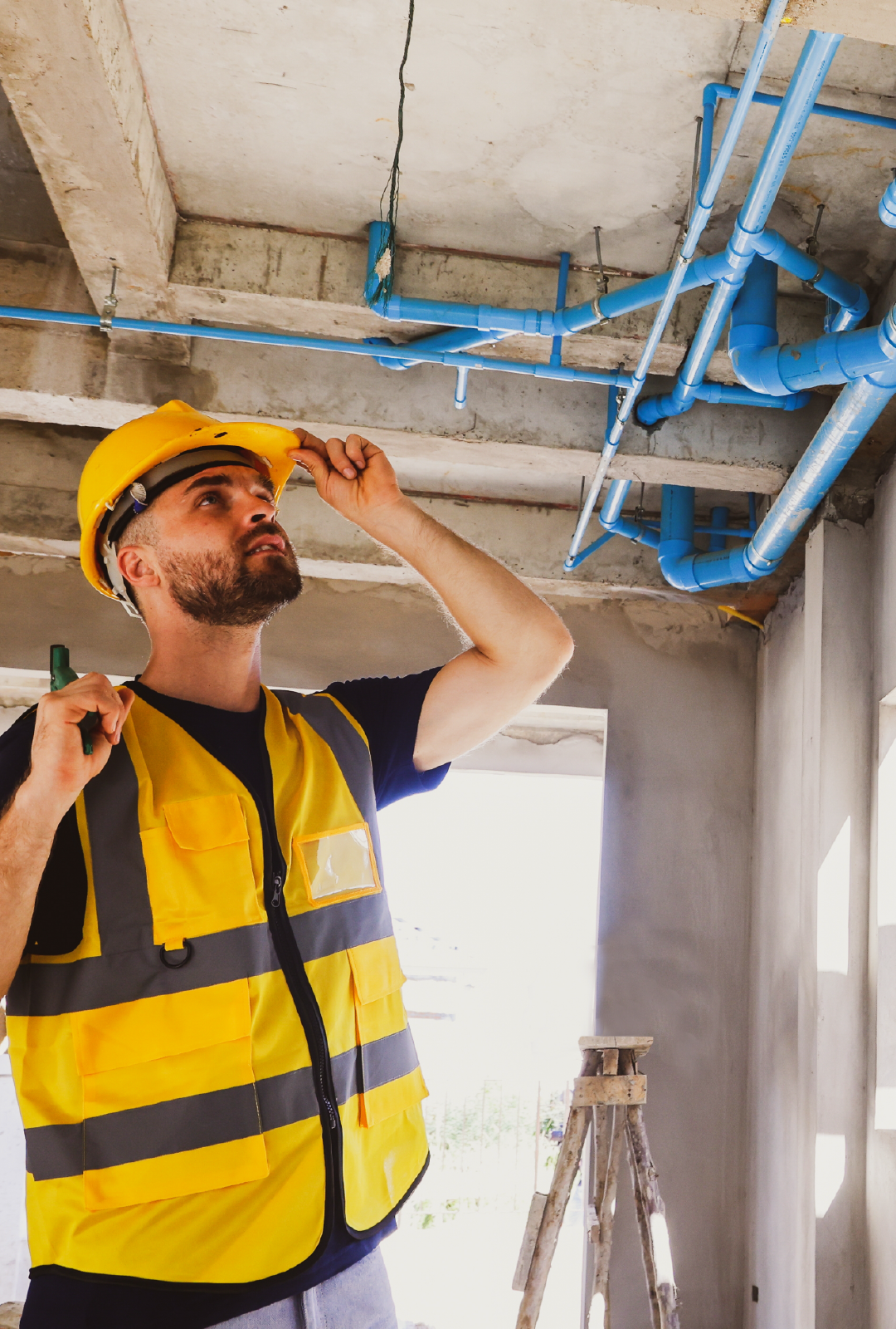 A man wearing a hard hat and safety vest is looking at pipes on the ceiling.