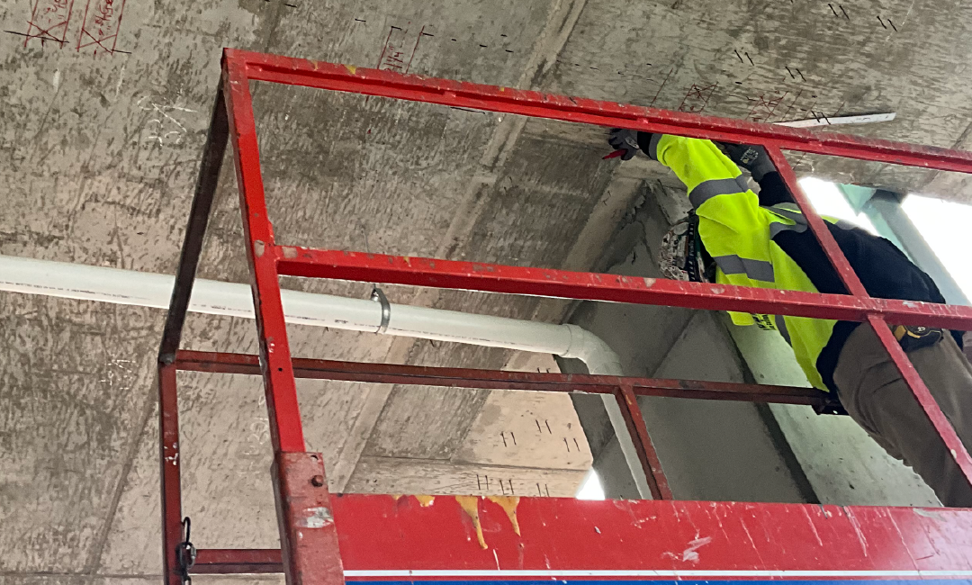 A man in a yellow vest is standing on a ladder looking up at the ceiling.