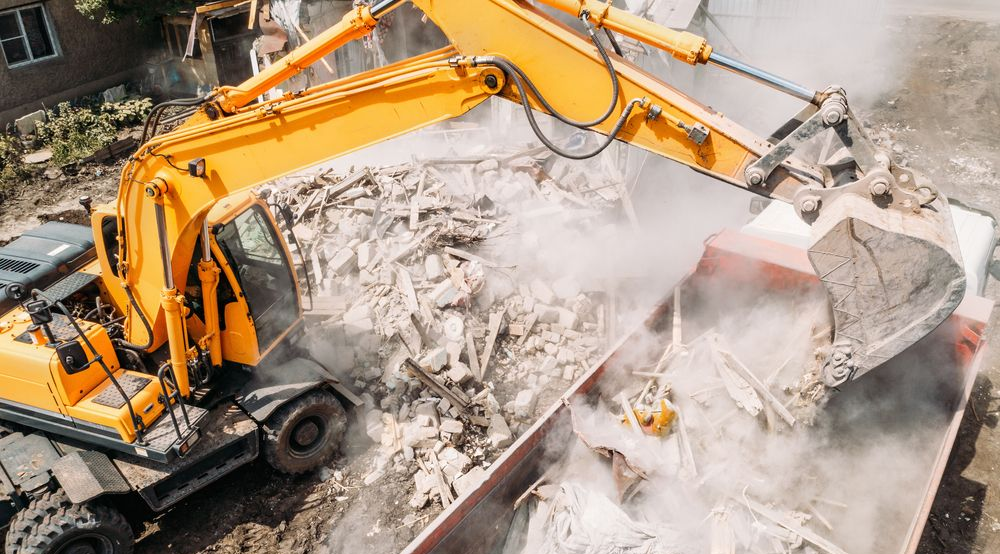 A yellow excavator is loading concrete into a dumpster.