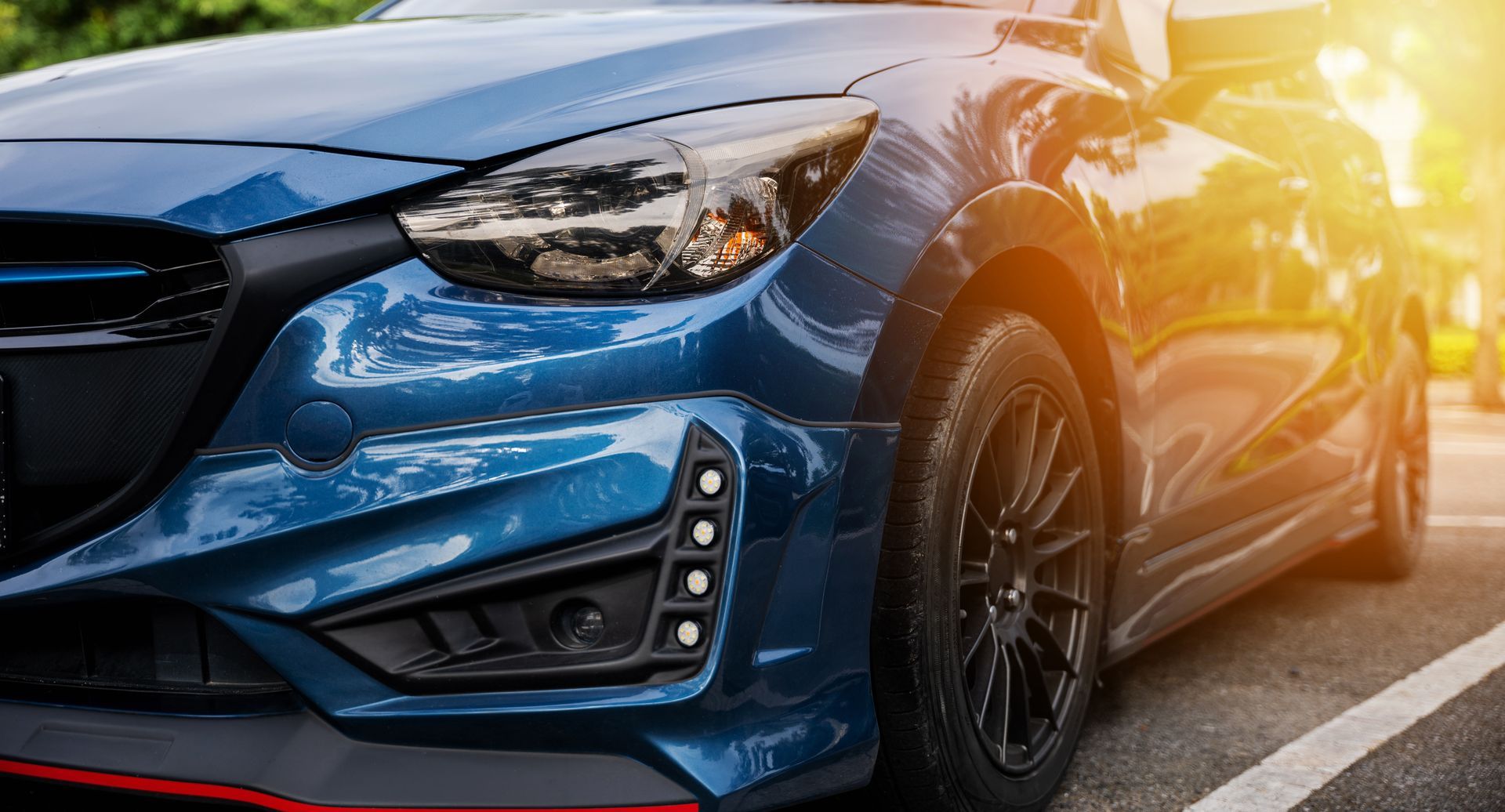 Blue car parked on a road, front view with sunlight reflecting off the hood and headlight.