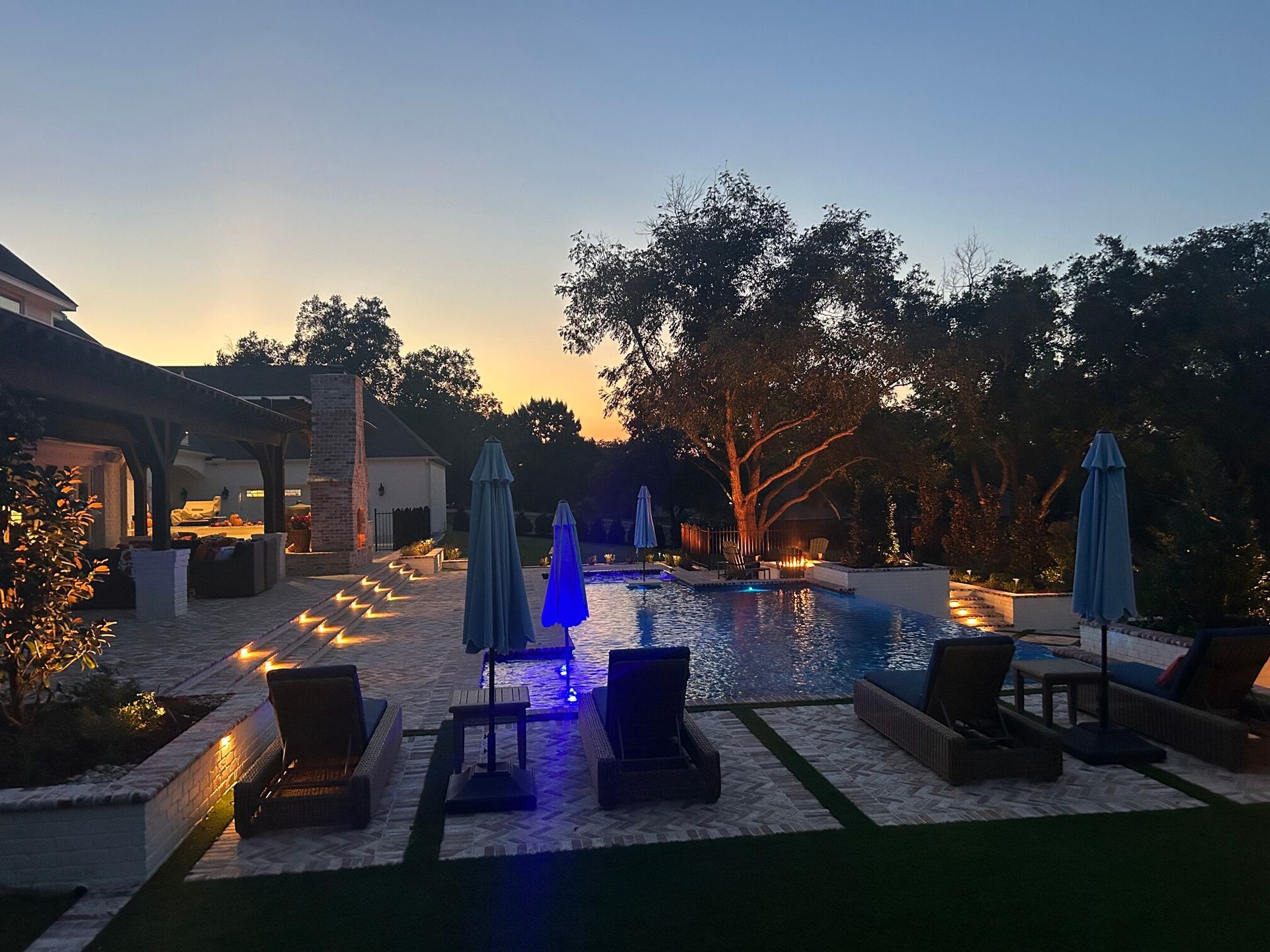 Poolside scene at dusk: pool, lounge chairs, umbrellas, trees, and string lights.