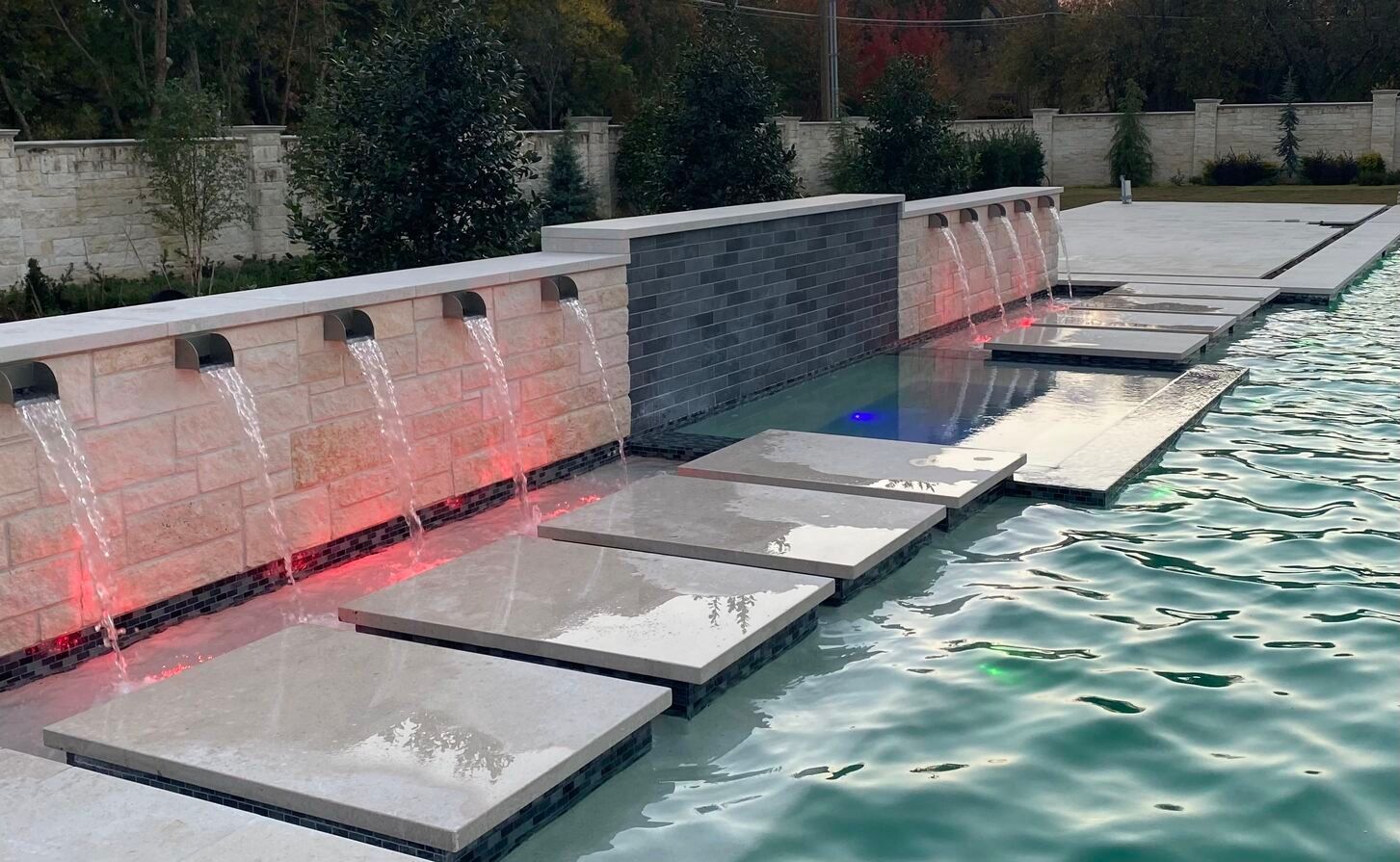 Water flows from metal spouts into a pool, with stepping stones. A stone wall is behind the spouts, and the water is lit with red lights.