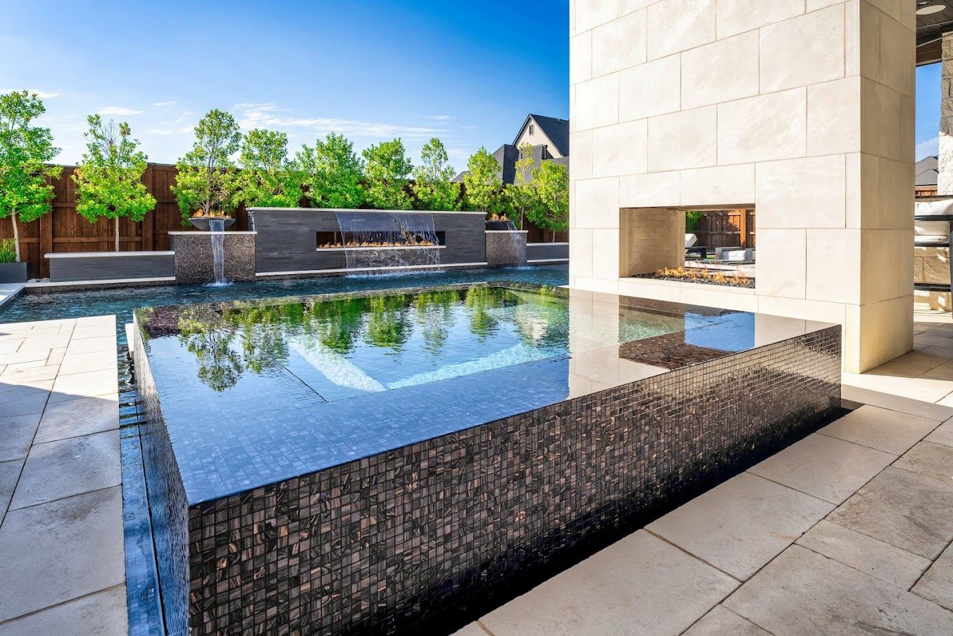 Outdoor pool with dark tile surround, reflecting sky, near stone fireplace and trees.