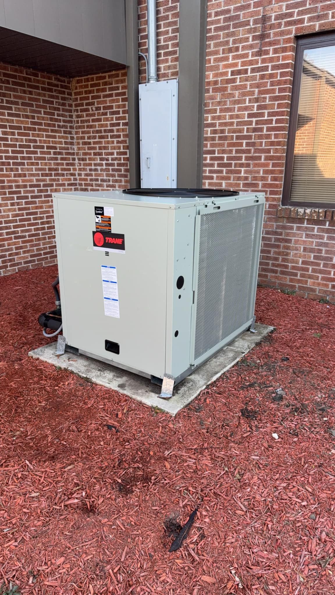 A large air conditioner is sitting on top of a pile of red gravel in front of a brick building.