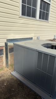 A large air conditioner is sitting outside of a house next to a window.