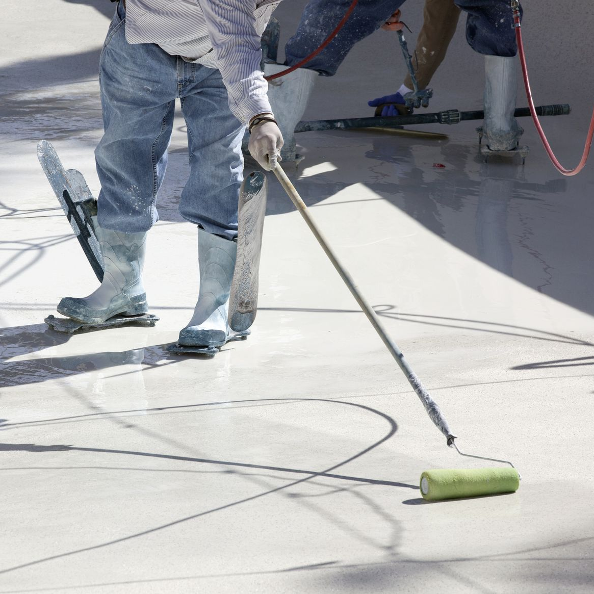 Workers applying coating to a concrete surface using rollers and other tools, outdoors.