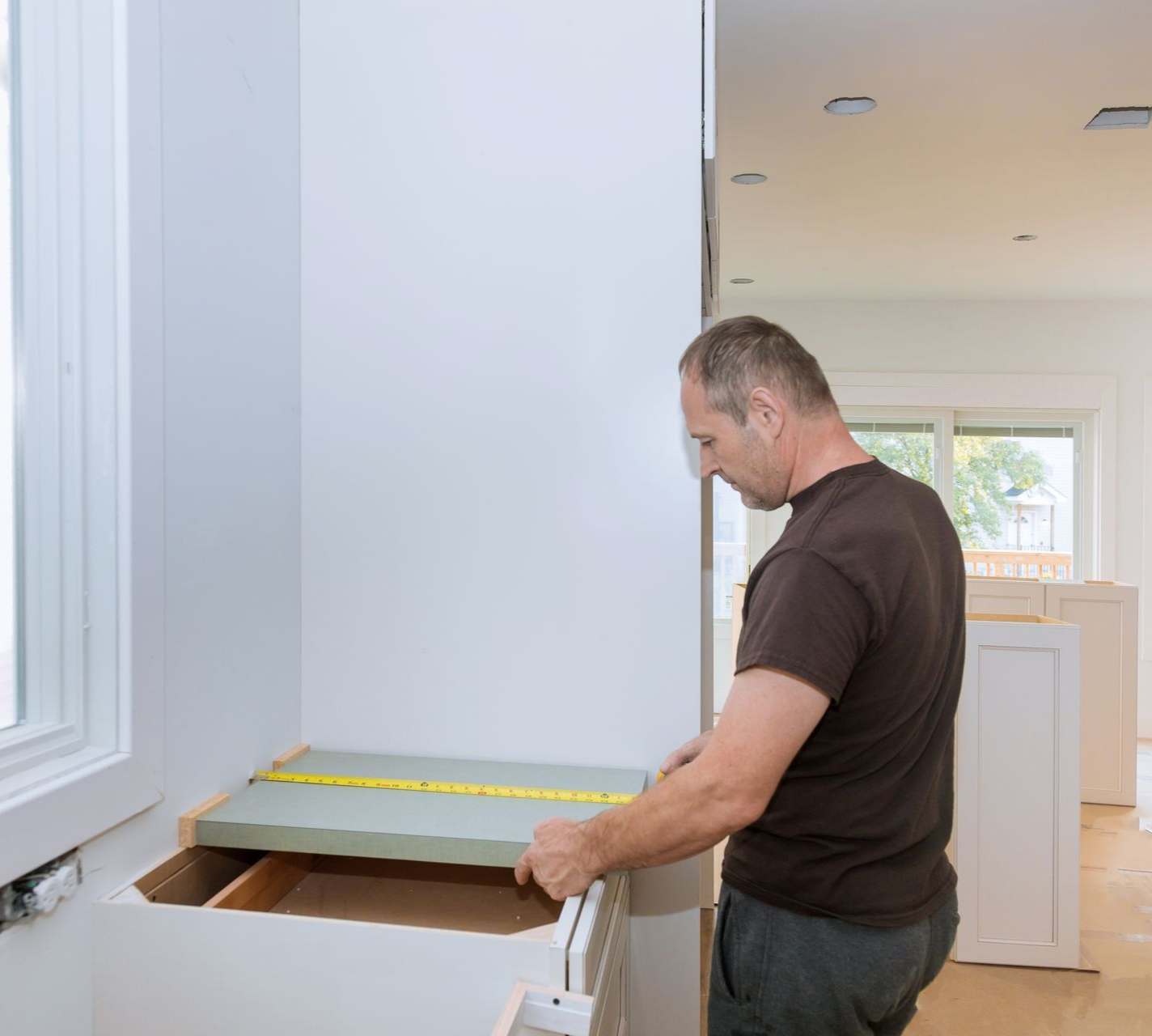 Man measures a countertop during kitchen renovation, standing near a window.