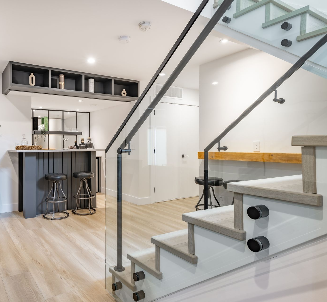 Modern interior with stairs, a bar, and built-in shelving, featuring light wood flooring and glass railings.