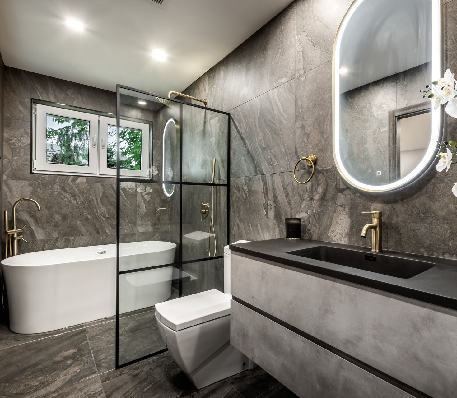Modern bathroom with gray stone walls, white soaking tub, glass shower, and vanity with black sink and gold fixtures.
