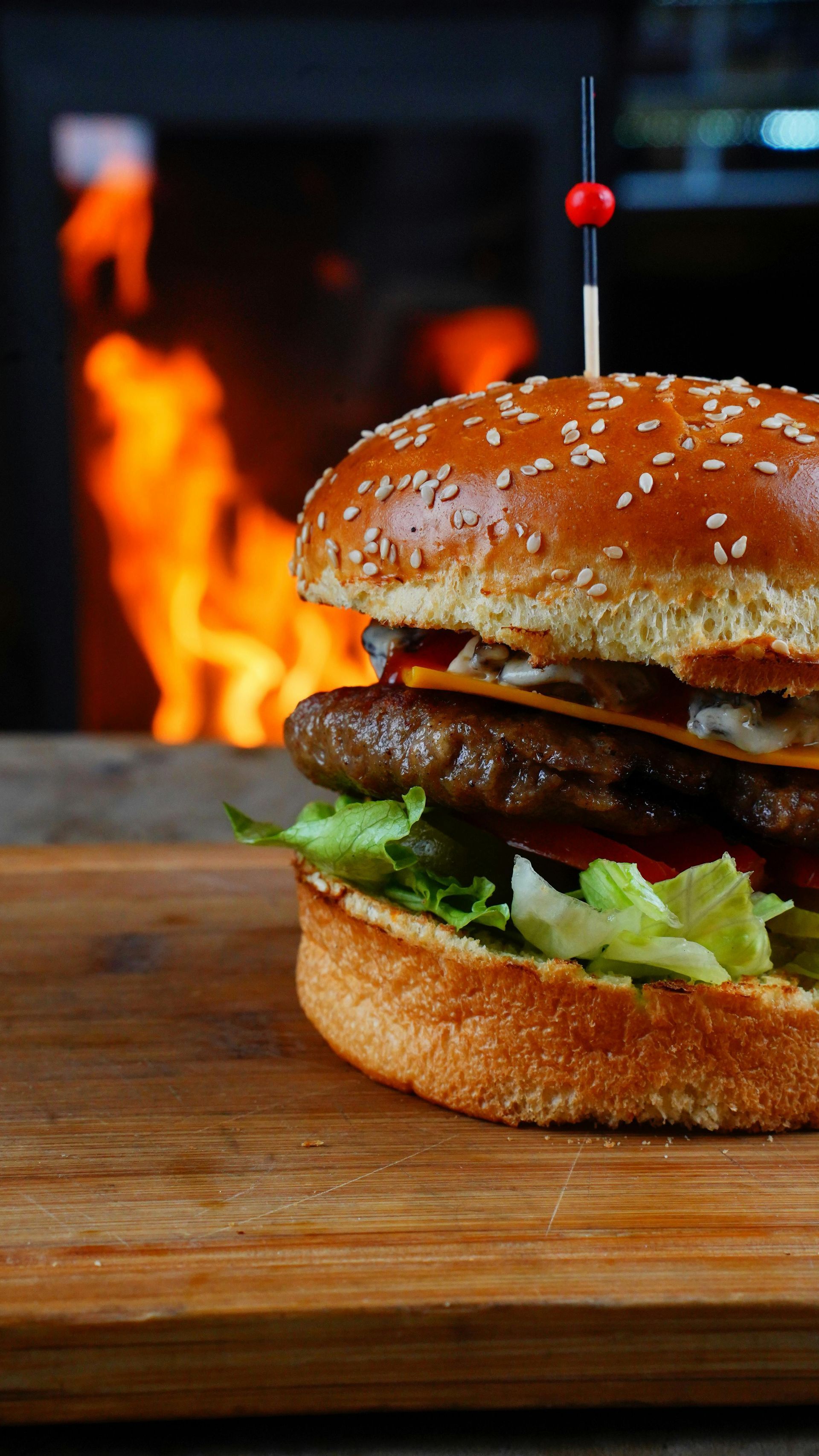 A sesame seed bun burger with lettuce and meat on a wooden board, set against a blurred, fiery fireplace background.
